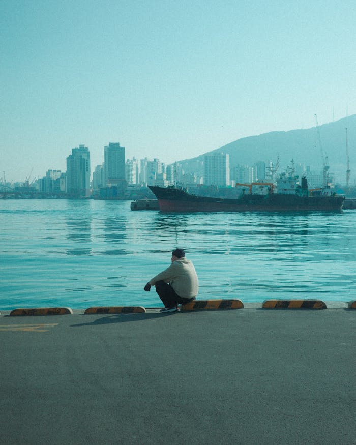 A man sits at the waterfront in Busan, South Korea, overlooking the harbor and city skyline.